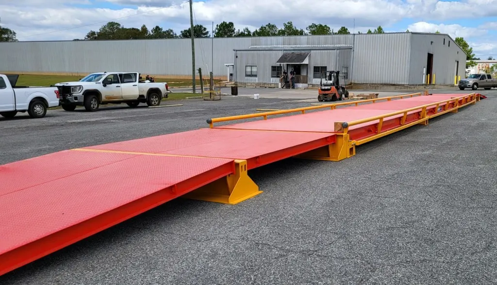 A Thurman Falcon 8565 PV portable truck scale on a paved yard, orange steel deck on yellow pedestal sub-frames with a steel ramp at the near end