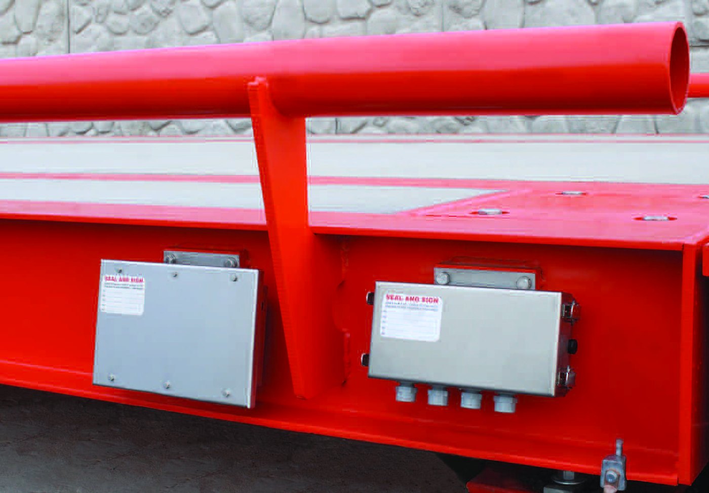Close-up of a Diamondback weighbridge: two sealed stainless-steel junction
enclosures bolted to the painted main beam, each carrying a seal-and-sign tag,
with the pipe rub rail above.