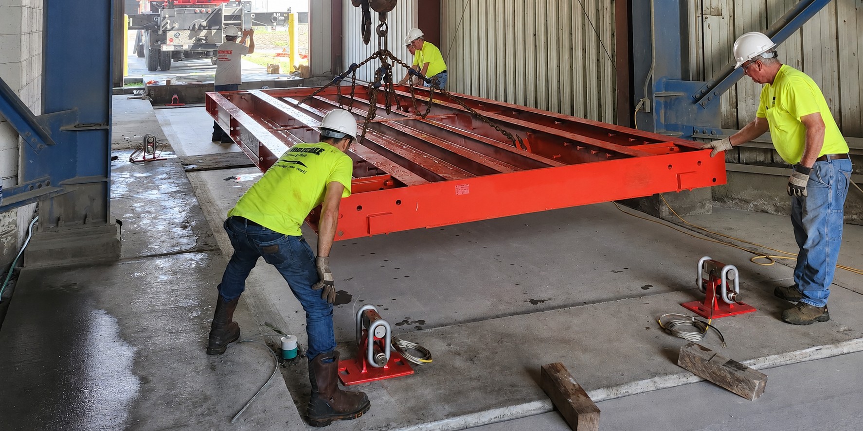 A Thurman crew in hard hats and high-visibility shirts guiding a suspended orange
Diamondback weighbridge module down onto its mounts inside a concrete batch plant, a
service truck crane holding the load.