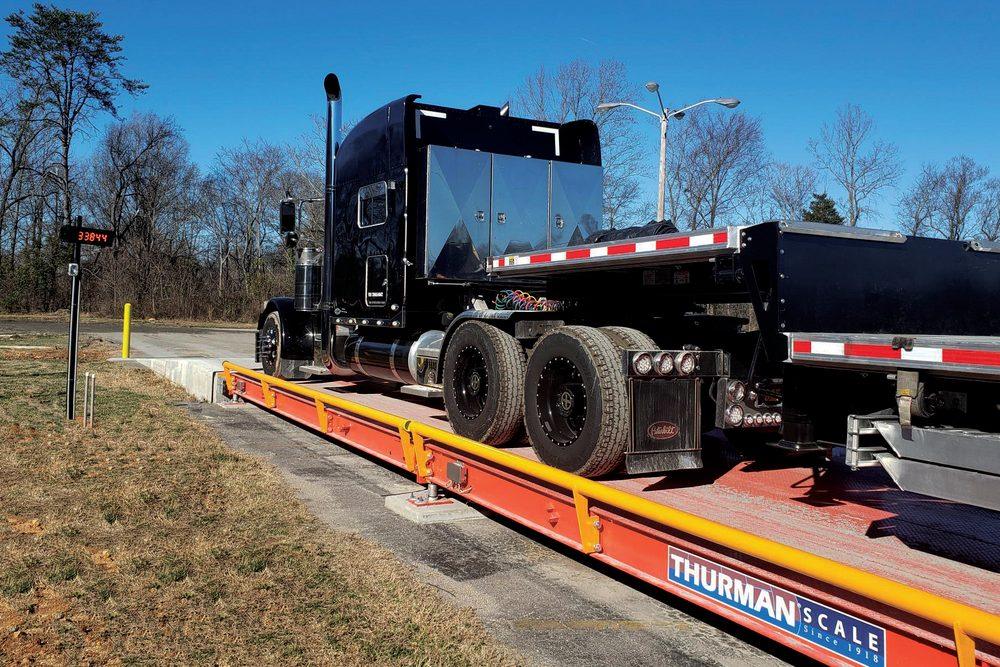 A Thurman Falcon 8565 above-ground steel deck truck scale in orange with yellow guide rails, a black tractor and dump trailer on the deck and a remote weight display at the approach