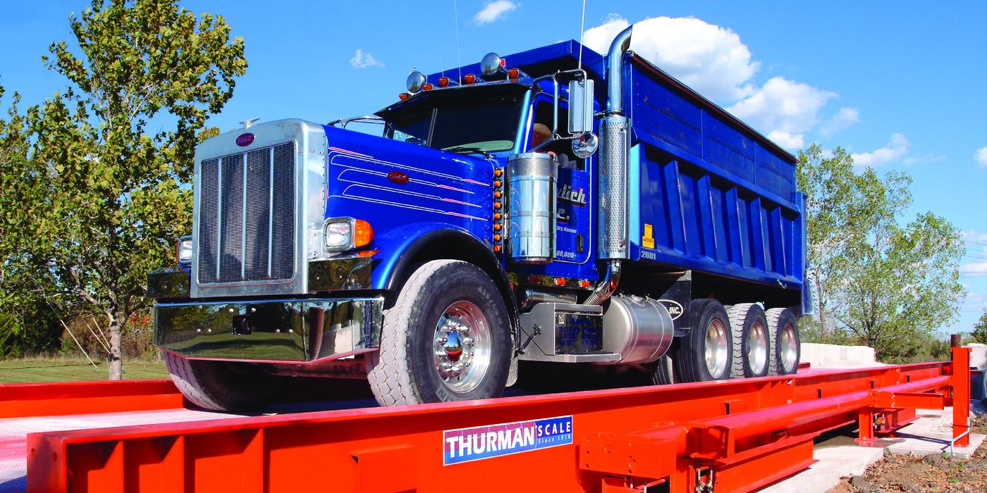 A loaded blue dump truck standing on an orange Thurman 8140 SAM side-arm truck scale, with the Thurman Scale plate on the weighbridge side rail