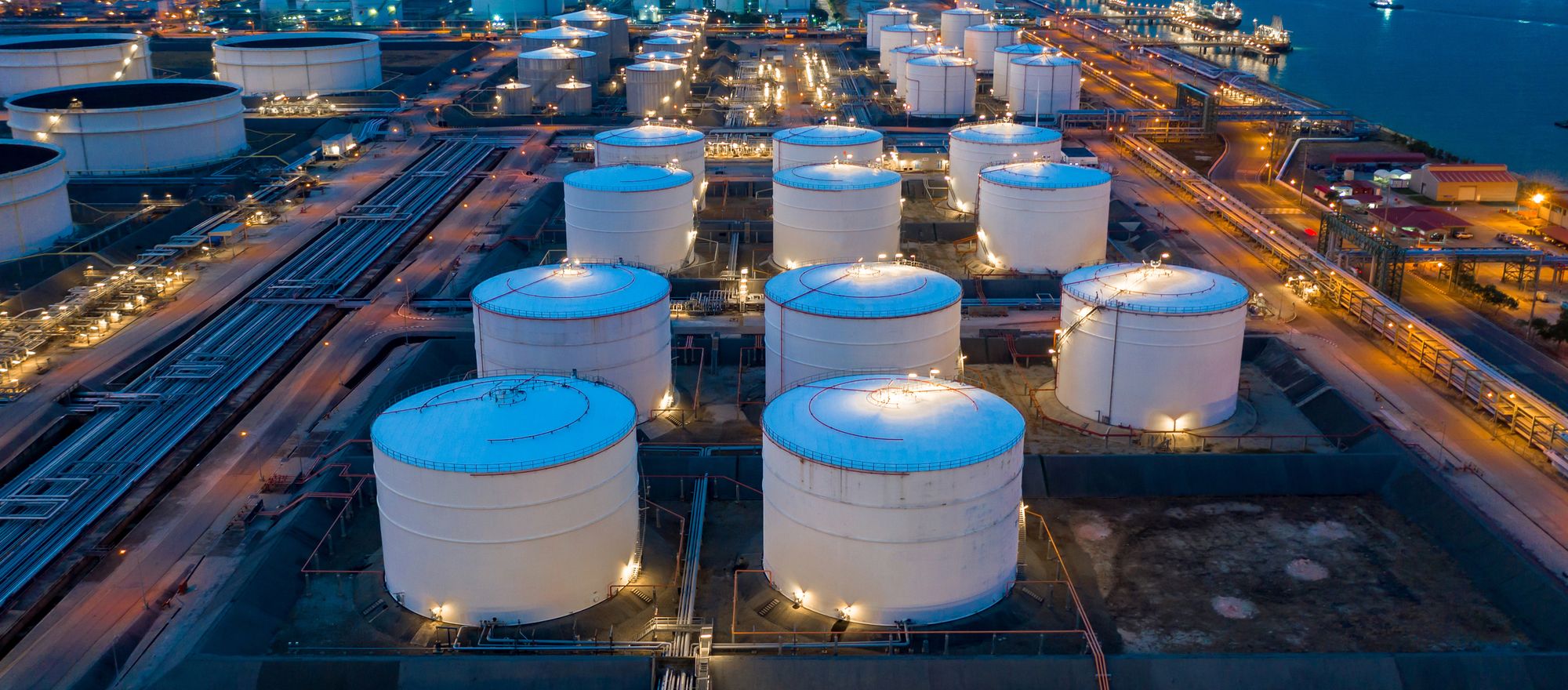 Aerial view of a tank farm at dusk, rows of white storage tanks lit along the pipe racks beside the water