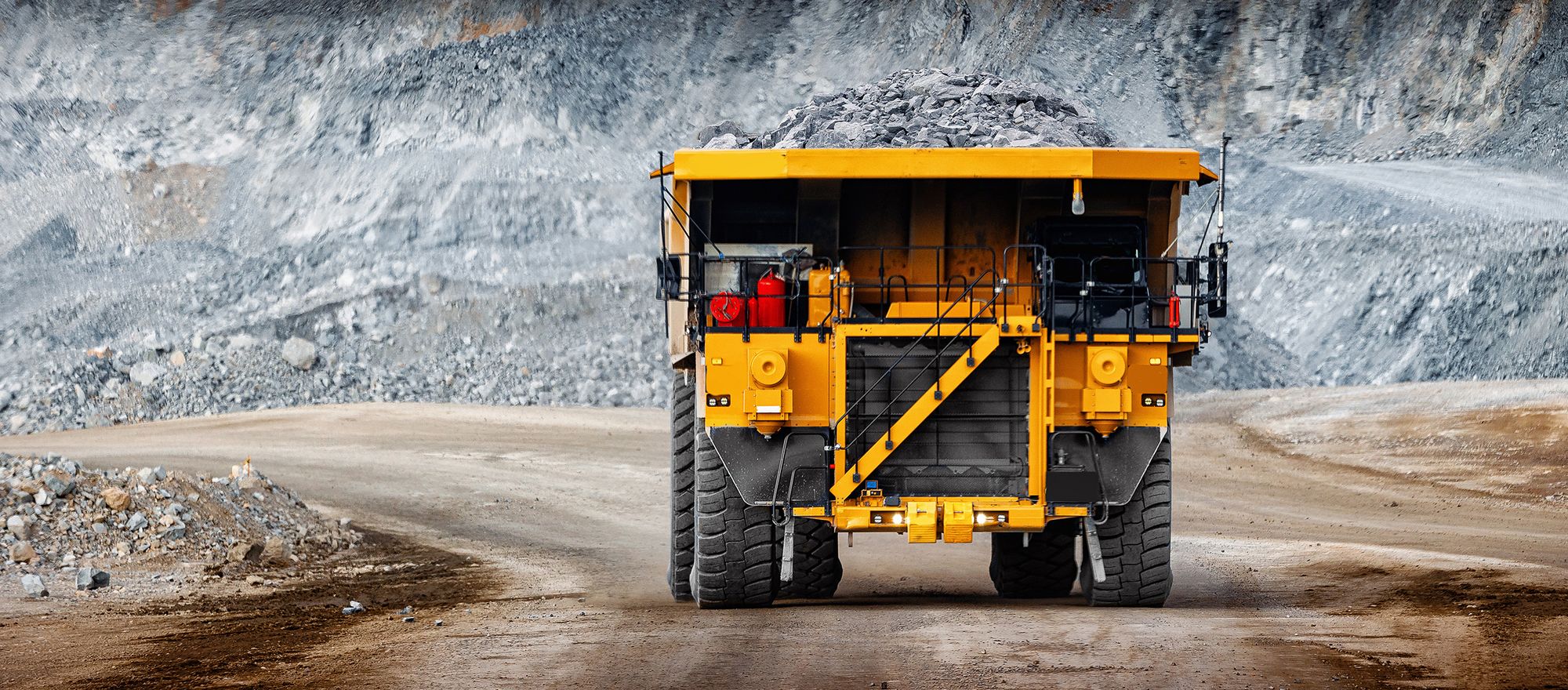 Loaded rigid-frame mining haul truck coming down a quarry haul road with a benched rock face behind it