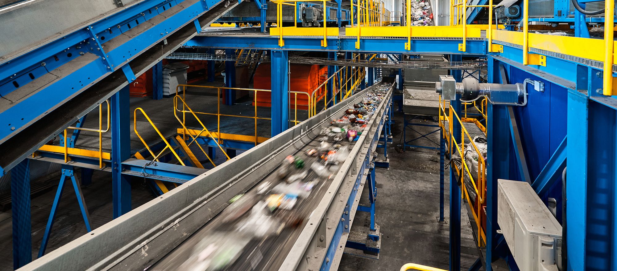 Mixed recyclables moving along a conveyor inside a materials recovery facility, under blue steel framing and yellow walkways
